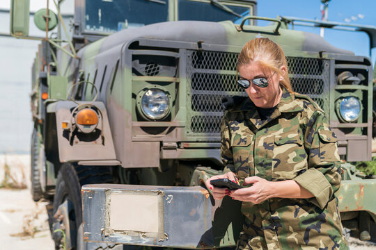 Mature blond female army soldier text messaging through smart phone while standing against truck at base on sunny day