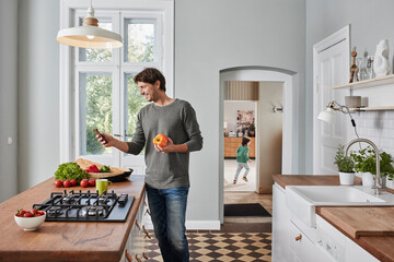Smiling man using smartphone and holding bell pepper in kitchen