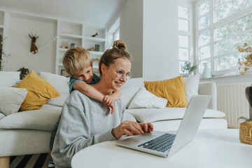 Woman working on laptop while boy hugging her from behind at home