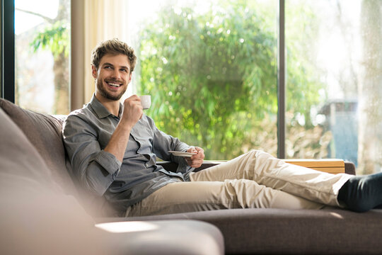 Laughing man relaxing on couch at home drinking coffee - Powered by Adobe