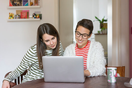 Two girlfriends at home sitting at table, using laptop