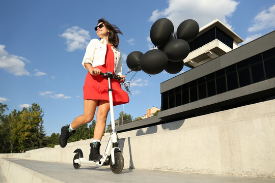 Young woman with black air balloons riding kick scooter along city street - Powered by Adobe
