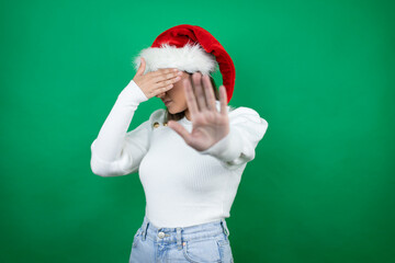 Young beautiful woman wearing a Santa hat and white sweater over green background covering eyes with hands and doing stop gesture with sad and fear expression. Embarrassed and negative concept.
