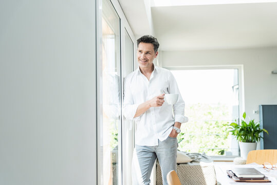 Mature man leaning on window, drinking coffee at home - Powered by Adobe