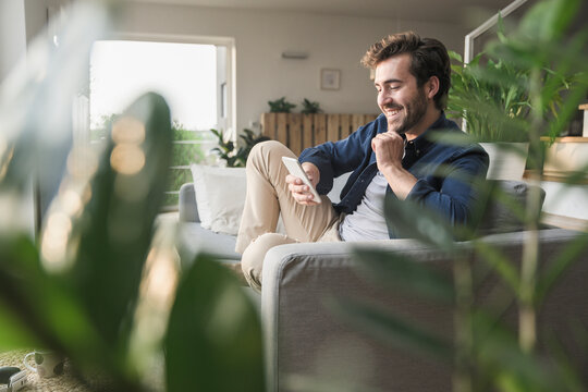 Young man sitting on couch at home, using smartphone