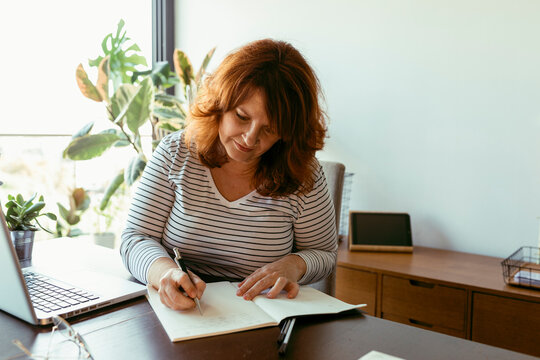 Woman writing in boo while using laptop on table at home