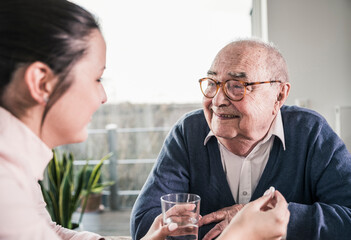 Woman holding pill and glass of water for smiling senior man