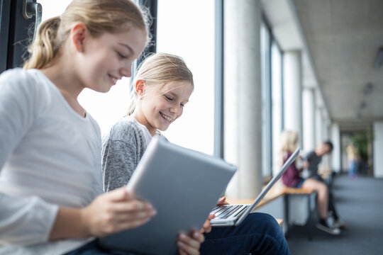 Happy schoolgirls with laptop and tablet on school corridor