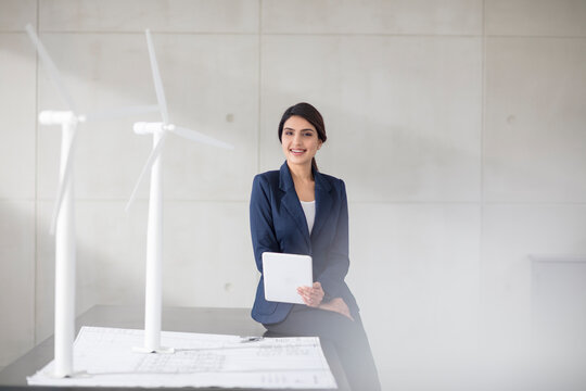 Portrait of smiling young woman with blueprint, wind turbine models and tablet in office