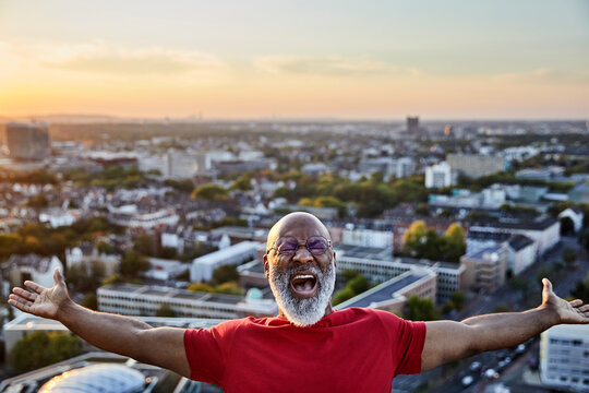 Bearded man shouting while standing with arms outstretched on building terrace in city during sunset