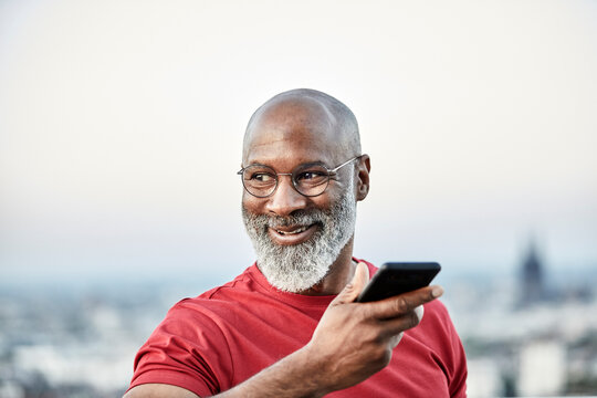 Smiling bald man talking through smart phone while looking away at building terrace during sunset