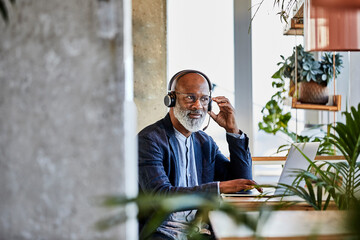 Mature businessman listening through headphones while sitting with laptop at table and working from home