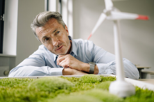 Businessman looking at wind turbine toy over moss frame while leaning on table at office