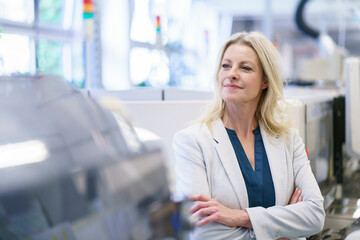 Thoughtful blond female professional standing with arms crossed in illuminated factory