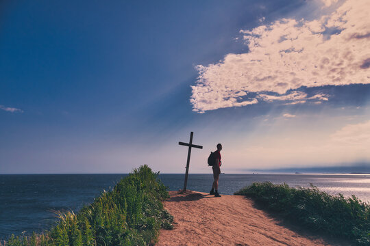 Hiker standing by summit cross while looking at sea against sky during sunset