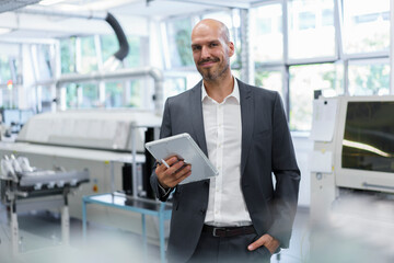 Smiling confident businessman holding digital tablet while standing with hand in pocket at bright factory