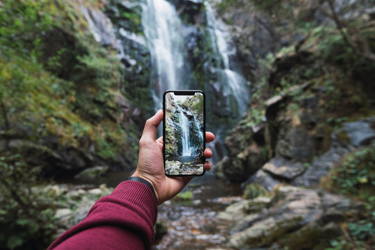 Hand of man holding smart phone while photographing famous Fervenza do Toxa in forest, Pontevedra Province, Galicia, Spain