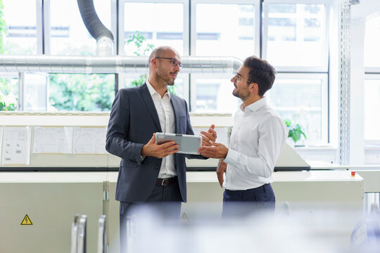 Male professionals discussing over digital tablet while standing against window at factory