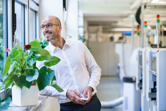 Thoughtful balding male technician looking away through window while holding remote control at factory