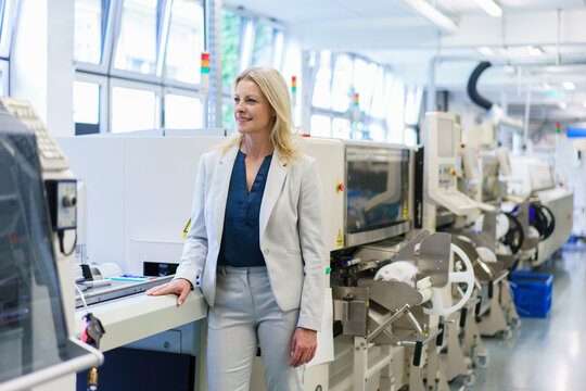 Thoughtful mature blond businesswoman standing by machinery while looking away at illuminated industry