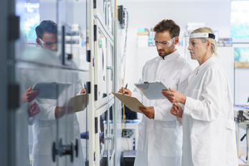 Young male scientist analyzing while writing on clipboard by female colleague holding digital tablet at laboratory