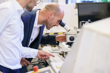 Young technician pointing to male colleague looking through microscope at laboratory