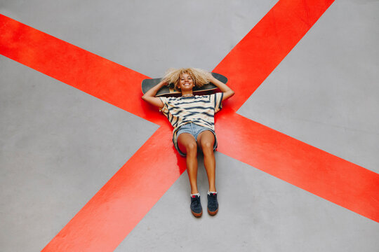 Smiling blond Afro woman lying with skateboard on orange road marking