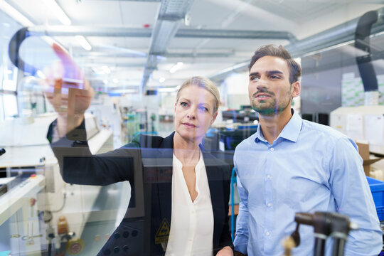Confident businesswoman planning with young engineer while pointing at glass interface in factory