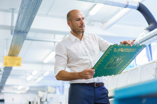 Confident balding male engineer looking at large circuit board while examining in illuminated factory