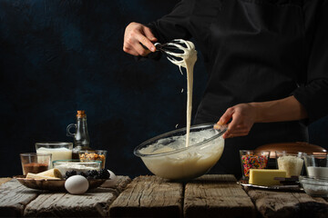 The professional chef makes dough for preparing waffles on rustic wooden table with ingredients on dark blue background. Backstage of cooking sweet dessert for breakfast. Frozen motion. Good recipe.