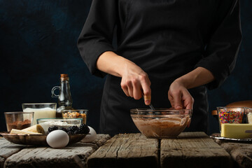 Fototapeta na wymiar The professional chef kneads dough for preparing waffles on rustic wooden table with ingredients on dark blue background. Backstage of cooking sweet dessert for breakfast. Frozen motion. Good recipe.