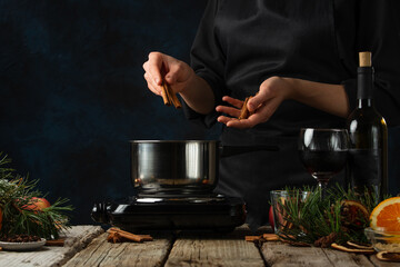 Chef pours cinnamon in pan for preparing mulled wine on rustic wooden table with festive composition background. Backstage of cooking hot drink with fragrant spices. Traditional recipe. Frozen motion