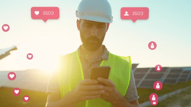 Engineer of solar power plant blogging on company page on social network. Young bearded man in yellow vest holding smartphone in hands, posting new content on background of solar panels.