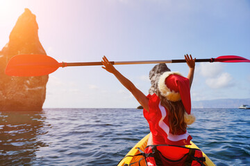 Young attractive brunette woman in red swimsuit and Santa hat, swimming on kayak around basalt rocks like in Iceland. Back view. Christmas holiday vacation and travel concept.