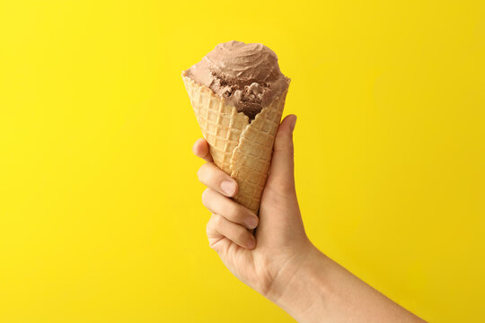 Woman holding waffle cone with delicious chocolate ice cream on yellow background, closeup