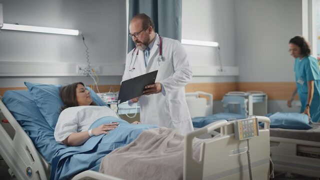 Hospital Ward: Friendly Latin Doctor Talks to Beautiful Female Patient Resting in Bed. Physician Shows Medical History Chart, Explains Test Results to Recovering after Successful Surgery Happy Woman