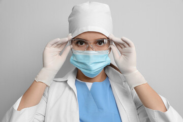 Doctor in protective mask and medical gloves putting on glasses against light grey background