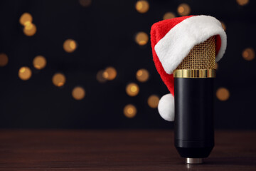 Microphone with Santa hat on wooden table against blurred lights, space for text. Christmas music