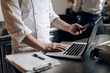 Crop chef using smartphone and laptop in kitchen