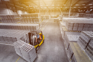 A forklift truck drives through a huge warehouse with stacks of vacuum-packed water or beer packages on pallets. View from above. Selective focus