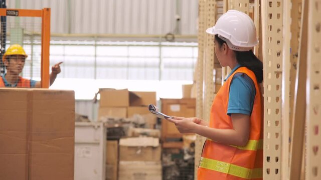 Asian woman warehouse manager hold clipboard for controlling stock and checking list inventory with worker using hand stacker in warehouse.
