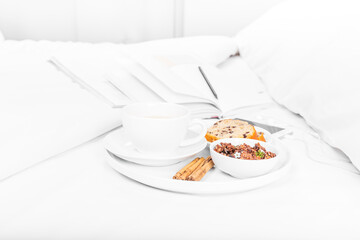 Breakfast tray on the bed, with tea, muffin, cinnamon and granola, white sheets, magazine, notebook, pencil, Smartphone, and earphones. White background.