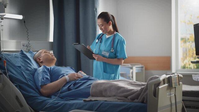 Hospital Ward: Friendly Chinese Head Nurse Fills Medical History Form, Talks to Male Patient Recovering in Bed. Beautiful Professional Nurse Helps Handsome Man Getting Better after Surgery. 