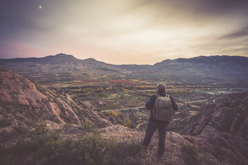 man with backpack and cap on the mountain enjoying the valley at sunset