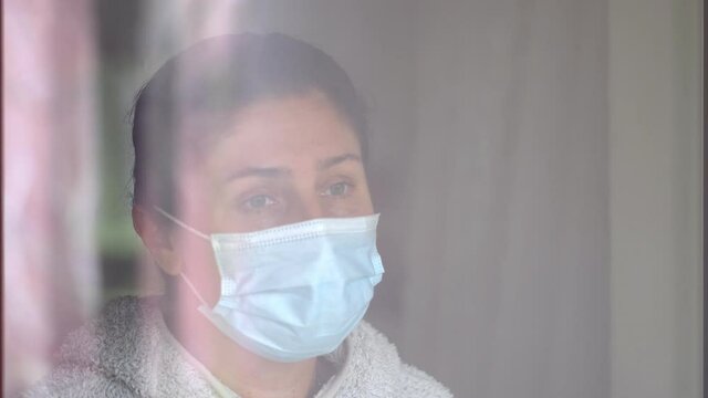 Young caucasian woman in a coronavirus mask touching with her hand to the glass of the window. Sad woman in quarantine. Pensive young woman in mask to protect herself from covid-19 while looking out