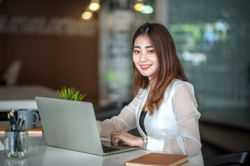 Beautiful young asian businesswoman is looking at camera and smiling happily while sitting working in office.