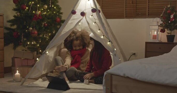 Two little girls having video call on tablet computer in decorated teepee tent at home celebrating Christmas with their friends online, holiday in the time of  COVID concepts