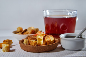 Square toasted pieces of homemade delicious rusk, hardtack, Dryasdust, zwieback, Liquid honey in a saucer and black tee in a cap on a white tablecloth.