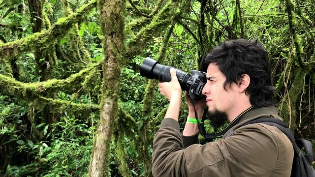 Young photographer man with black hair and beard, and brown jacket taking pictures with a camera an telephoto in the middle of a green forest with moss and lianas.