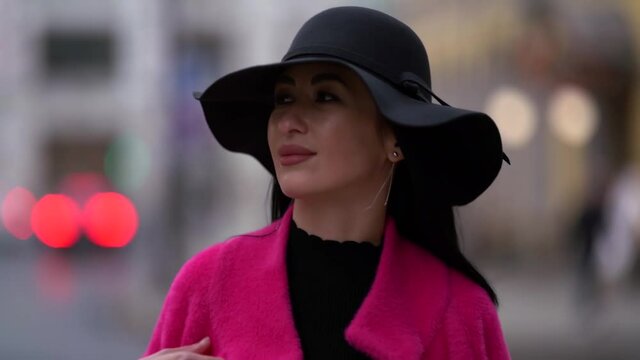 Close-up portrait of a stylish and confident black-haired woman in a pink coat and black hat walking and posing on a blurry background of a busy city street.